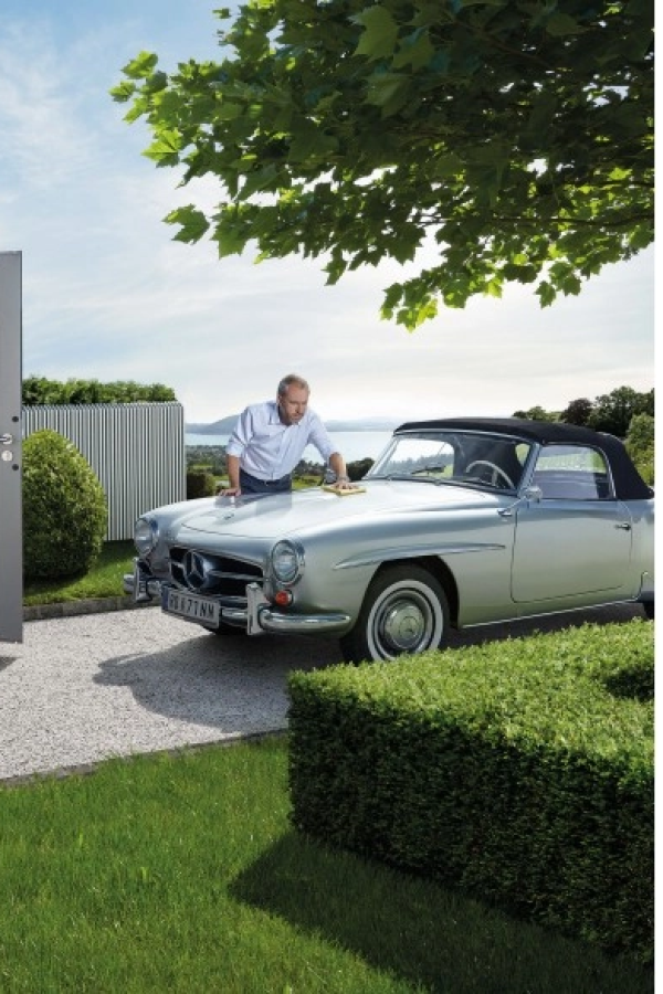Homme essuie capot Mercedes convertible argent devant haie, gravier et paysage lac