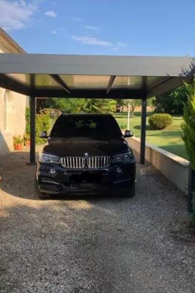 Carport alu anthracite avec toit clair abritant SUV noir sur gravier, jardin et ciel bleu