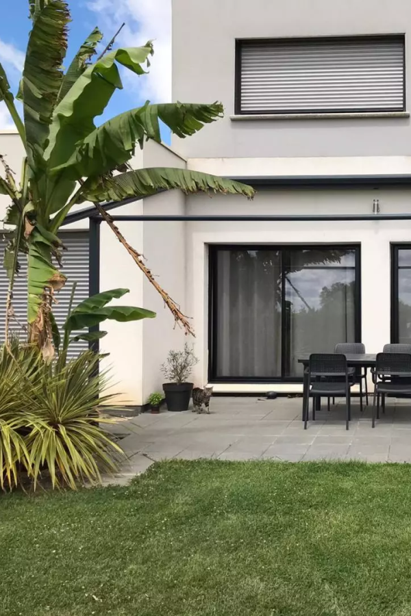 Maison claire avec baies noires, volet roulant gris à l'étage, terrasse carrelée et bananier