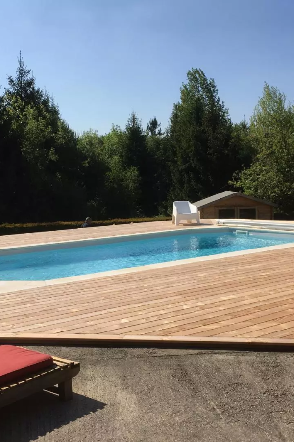 Piscine rectangulaire bleue encastrée dans grande terrasse bois, jardin boisé et cabane