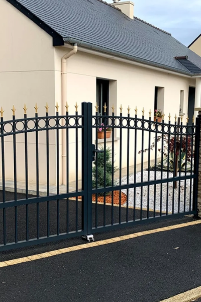 Portail battant noir à barreaux avec finitions dorées devant maison beige toit ardoise