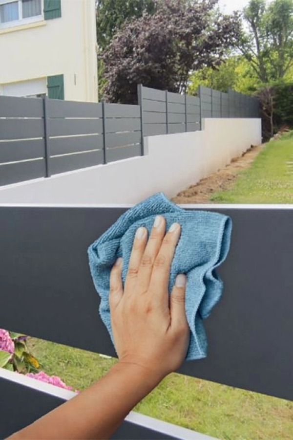 Main nettoie clôture anthracite à lames avec chiffon microfibre bleu, jardin en fond