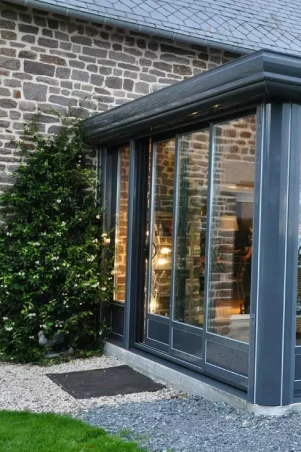 Véranda gris anthracite adossée à mur en pierre, lierre fleuri et gravier devant l'entrée