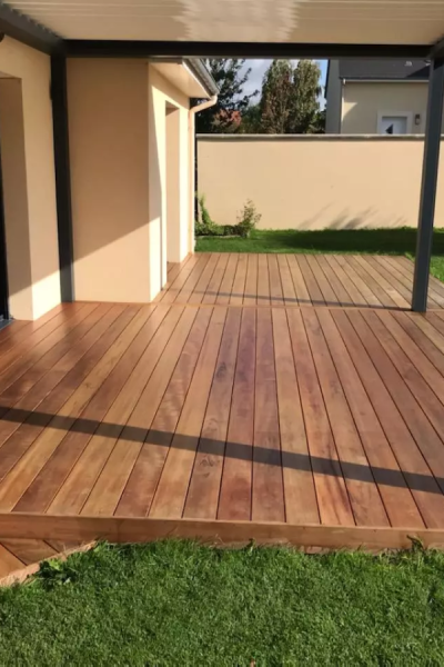 Terrasse bois exotique brun sous pergola sombre, pelouse et mur beige en contrebas