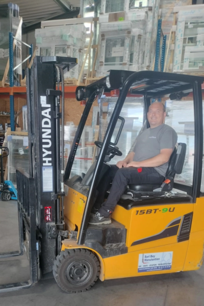 Homme souriant dans un chariot élévateur Hyundai devant racks de menuiseries en entrepôt