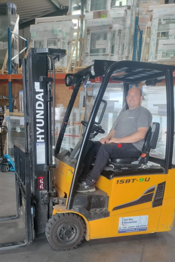 Homme souriant dans un chariot élévateur Hyundai devant racks de menuiseries en entrepôt