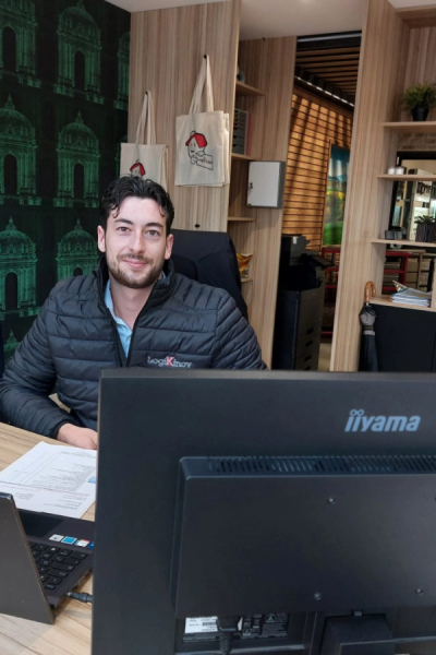 Homme en doudoune noire logo brodé à un bureau, sacs tote et papier peint vert