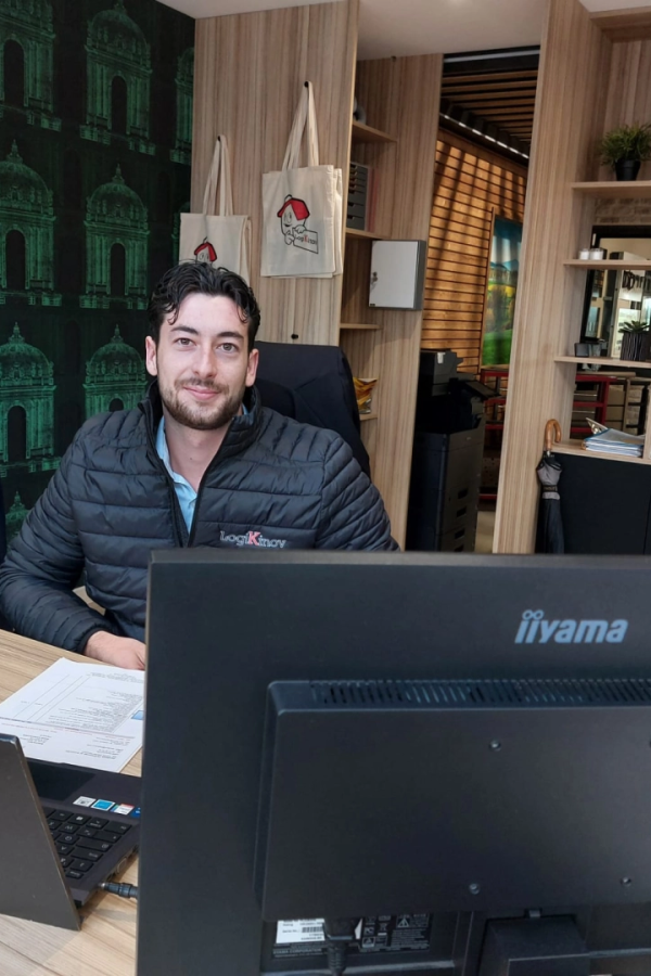 Homme en doudoune noire logo brodé à un bureau, sacs tote et papier peint vert