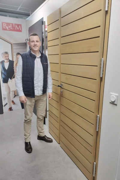 Homme souriant devant porte d'entrée bois clair à lames horizontales dans showroom Bel'M