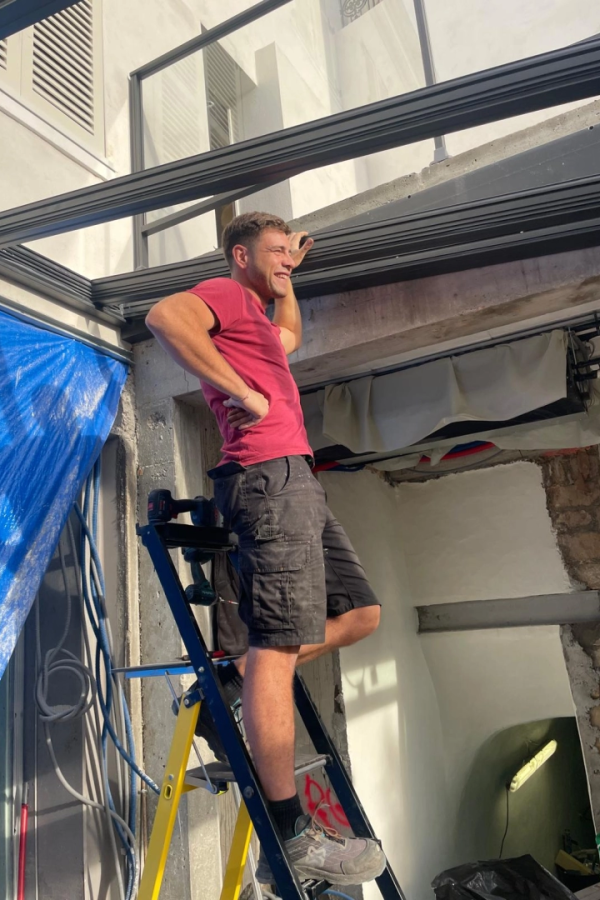 Chef d'équipe souriant sur échelle devant ossature noire de véranda en chantier