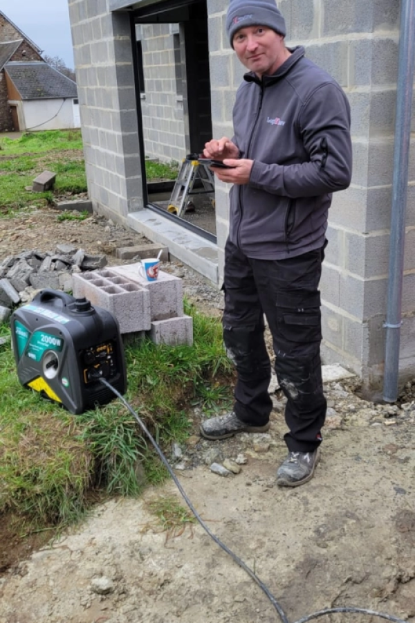 Poseur en tenue grise avec smartphone à côté d'un groupe électrogène 2000 W sur chantier parpaings