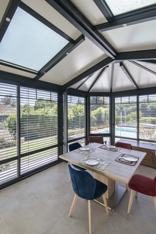 Intérieur véranda anthracite, stores horizontaux, table à manger bleue et vue jardin piscine