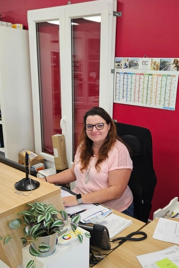 Femme à lunettes souriante à un bureau devant mur rouge et planning affiché à la fenêtre