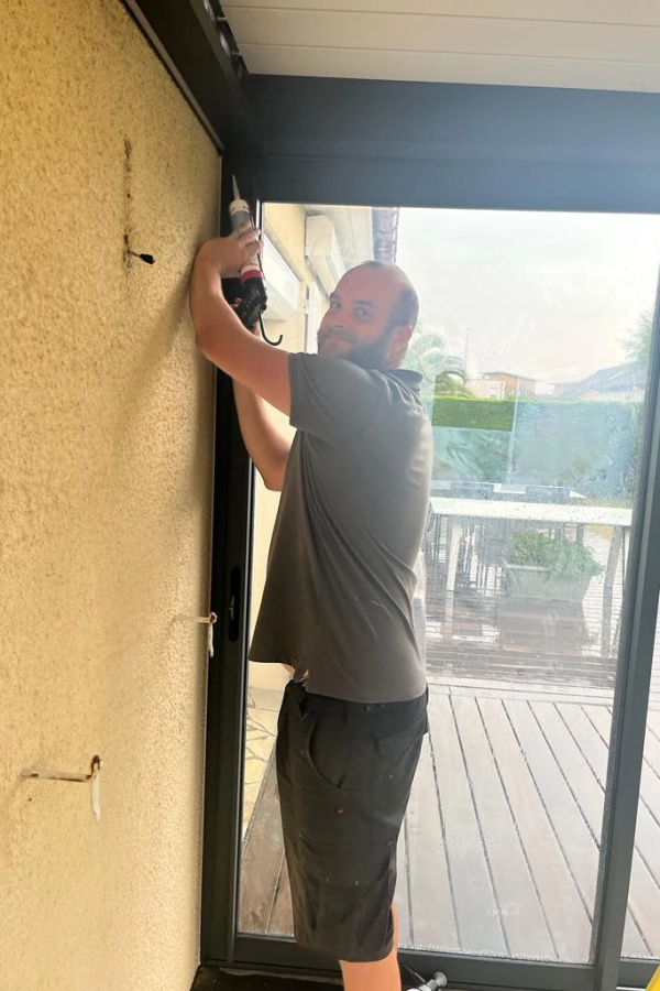 Technicien applique mastic blanc sur cadre anthracite d'une baie, terrasse bois visible