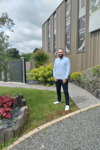 Technicien souriant devant agence avec affiches portail, volet et clôture et jardin paysagé