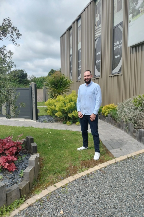 Technicien souriant devant agence avec affiches portail, volet et clôture et jardin paysagé