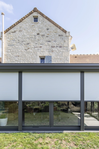 Volet roulant blanc mi-hauteur sur extension vitrée anthracite contre façade en pierre