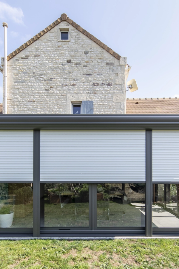 Volet roulant blanc mi-hauteur sur extension vitrée anthracite contre façade en pierre