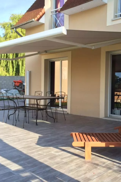 Store banne taupe déployé sur terrasse carrelée effet bois, table métal et transat devant maison