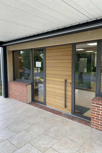 Porte d'entrée en lames bois clair avec poignée noire, vitrines anthracite et pavés travertine