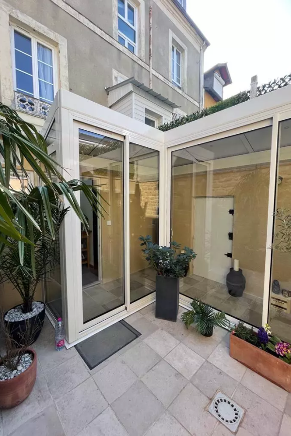 Véranda angle alu beige vitrée sur terrasse dalles, pots plantes devant maison pierre