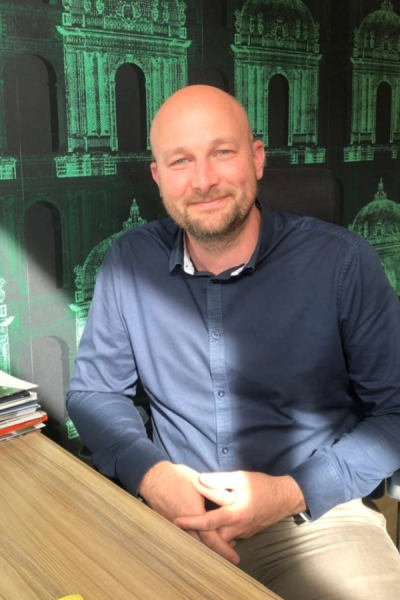 Homme chauve barbe souriant assis à un bureau, lumière naturelle et papier peint vert