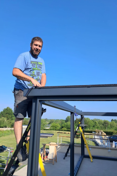 Installateur sur échelle Centaure posant poutre anthracite de pergola sur terrasse ensoleillée