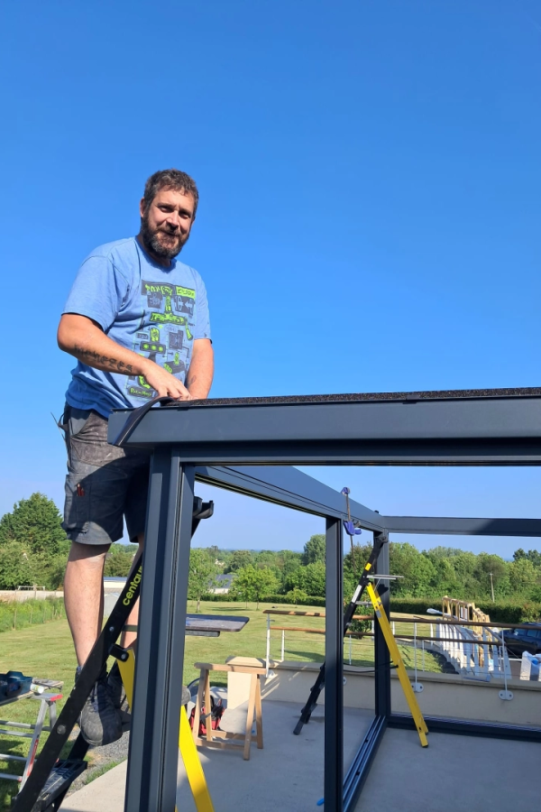 Installateur sur échelle Centaure posant poutre anthracite de pergola sur terrasse ensoleillée