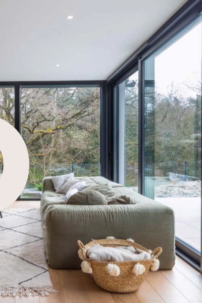 Salon avec grande baie coulissante alu noire ouvrant sur forêt, canapé vert et parquet