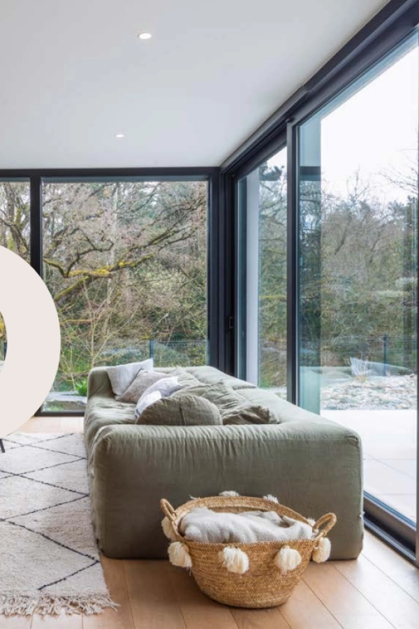 Salon avec grande baie coulissante alu noire ouvrant sur forêt, canapé vert et parquet