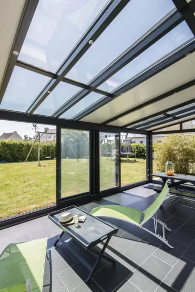 Intérieur véranda lumineux, cadre noir, chaises vertes et table, vue jardin et terrasse ardoise
