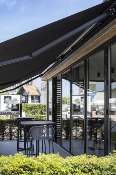 Store banne noir déployé sur terrasse du bistro L'Échiquier avec mobilier et haie verte