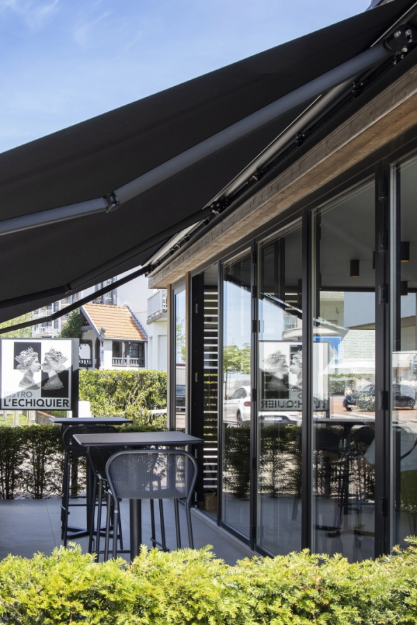 Store banne noir déployé sur terrasse du bistro L'Échiquier avec mobilier et haie verte