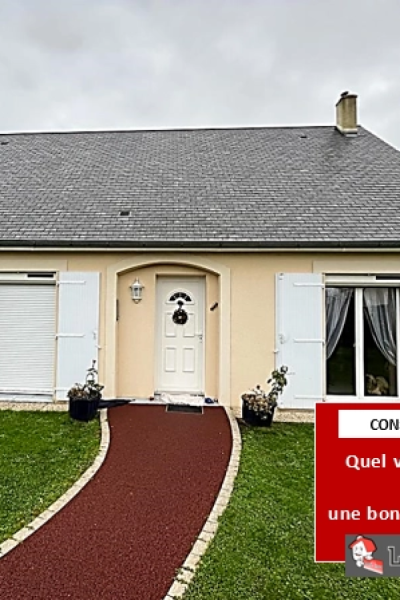 Maison beige toiture ardoise, volets blancs, allée rouge bordée de pavés et pelouse