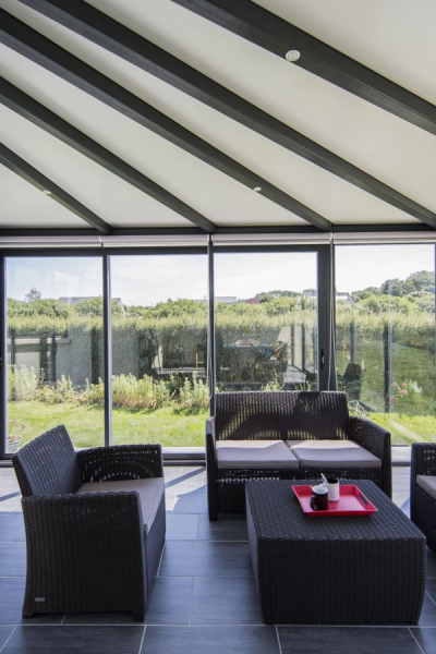 Intérieur véranda avec salon resine tressée grise, carrelage foncé et vue jardin par baies vitrées