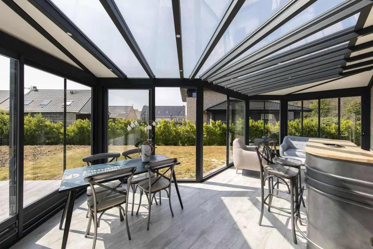 Véranda noire vitrée avec table, fauteuils et baril transformé en comptoir, vue jardin