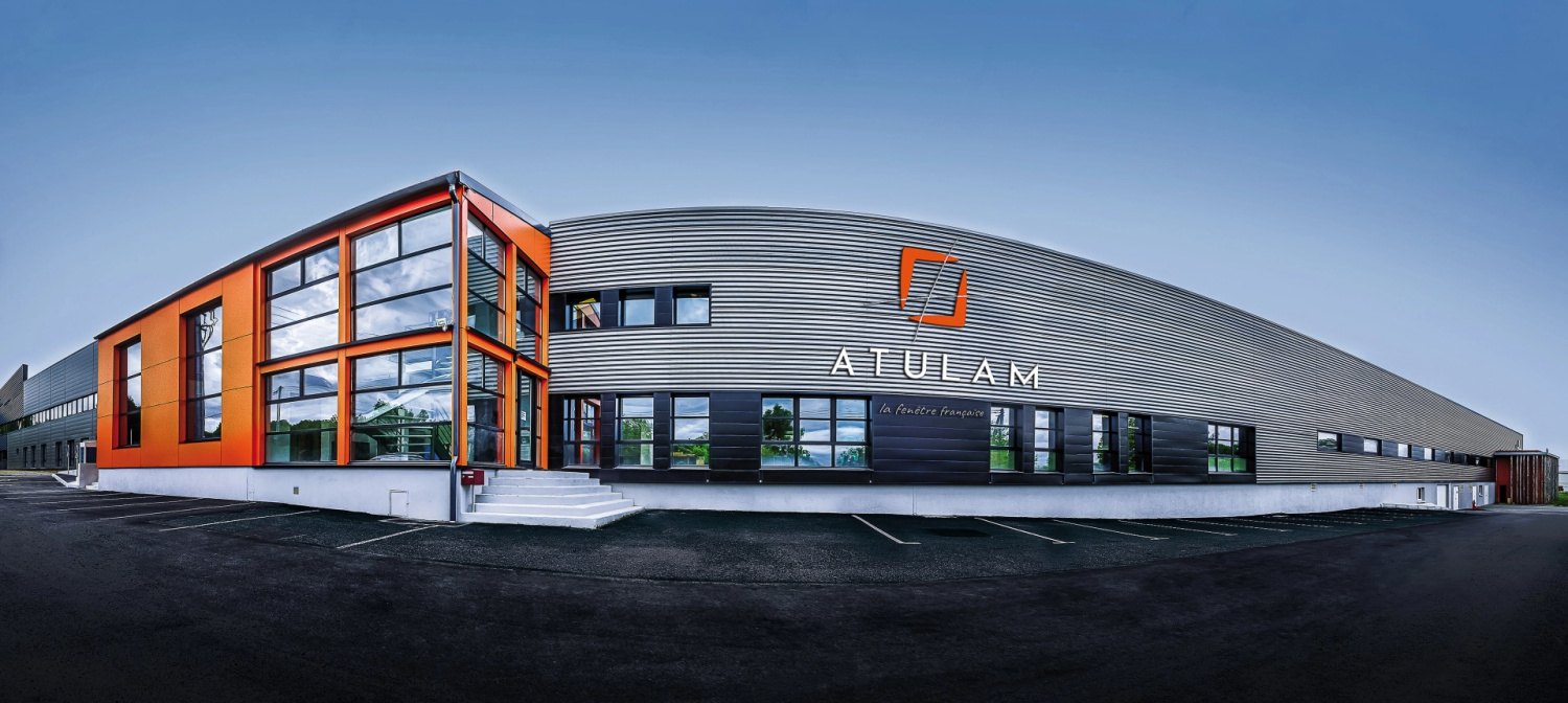 Façade grise usine Atulam avec logo orange la fenêtre française et verrière orange