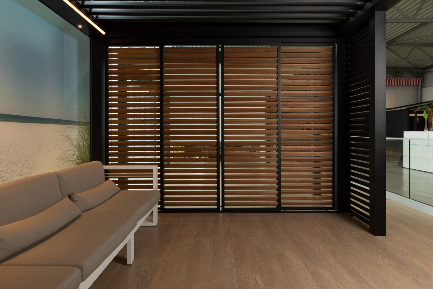 Showroom pergola noire LED, paroi persiennes bois marron et canapé gris, fresque plage
