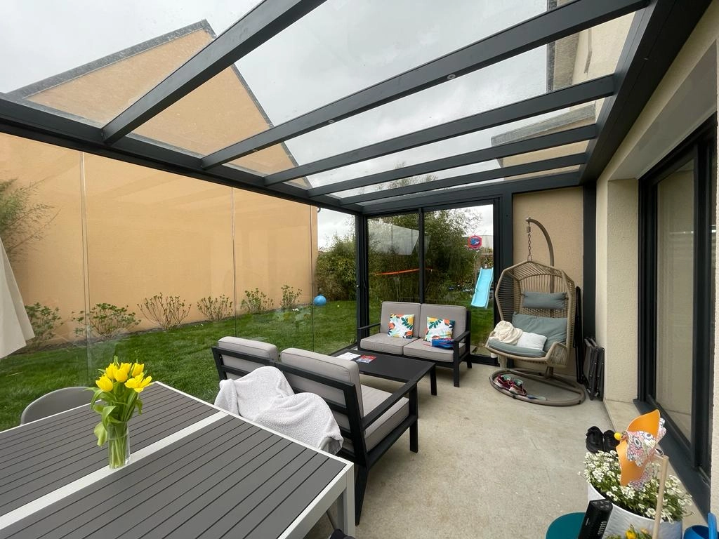 Pergola vitrée anthracite de nuit sur terrasse, salon, table et fauteuil suspendu
