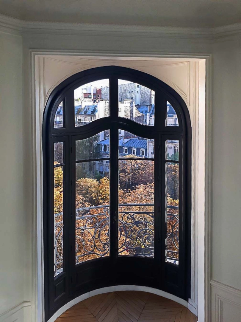 Fenêtre cintrée noire en alcôve, parquet chevrons et vue arbres automnaux et toits ville