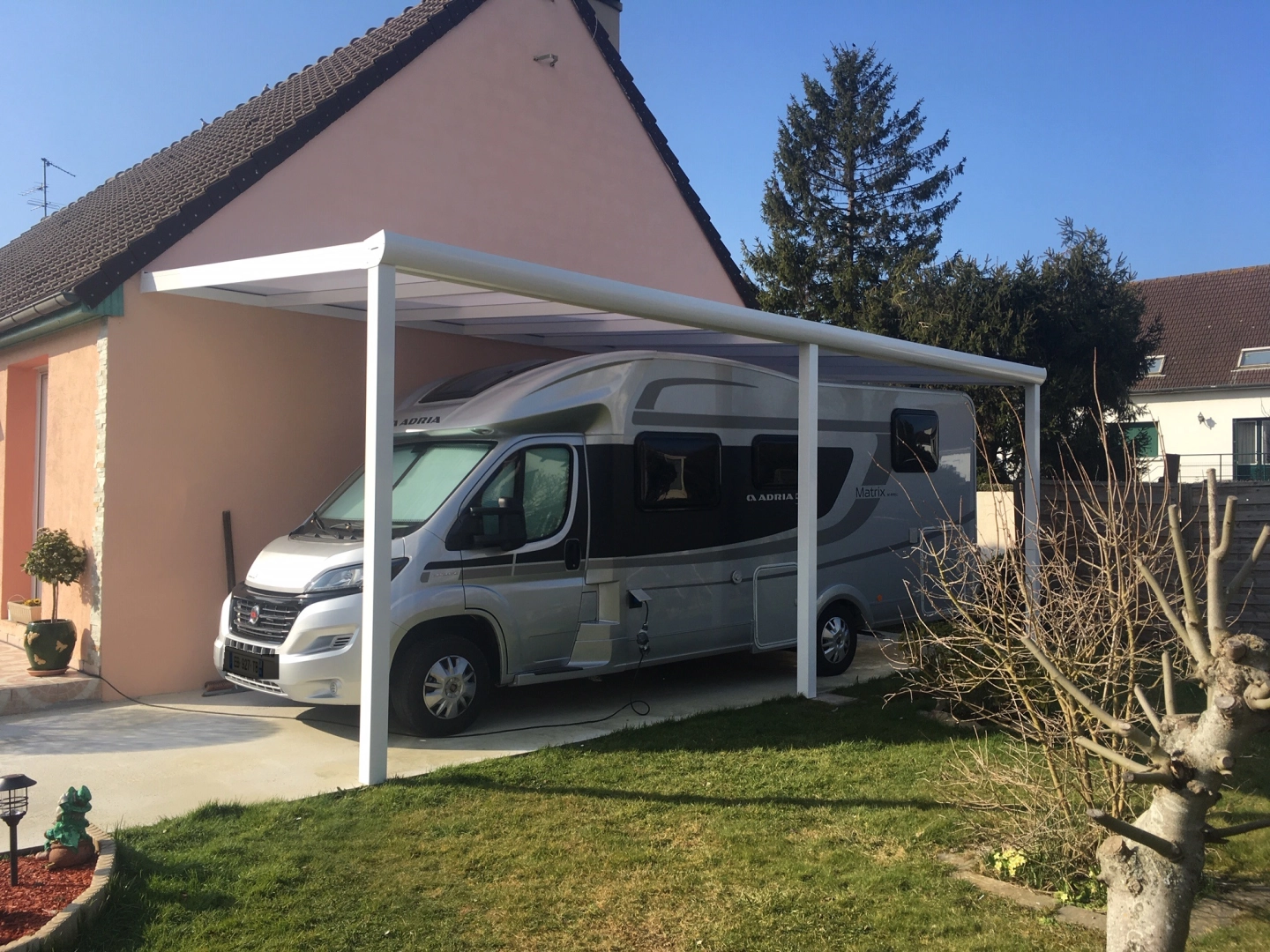 Camping-car gris Adria sous carport adossé blanc devant maison rose et pelouse