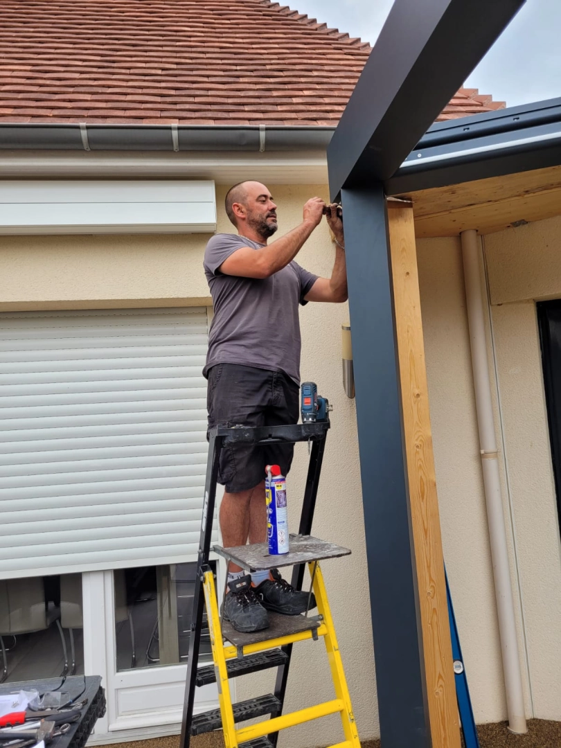 Installateur sur escabeau fixant poteau pergola aluminium gris contre maison beige