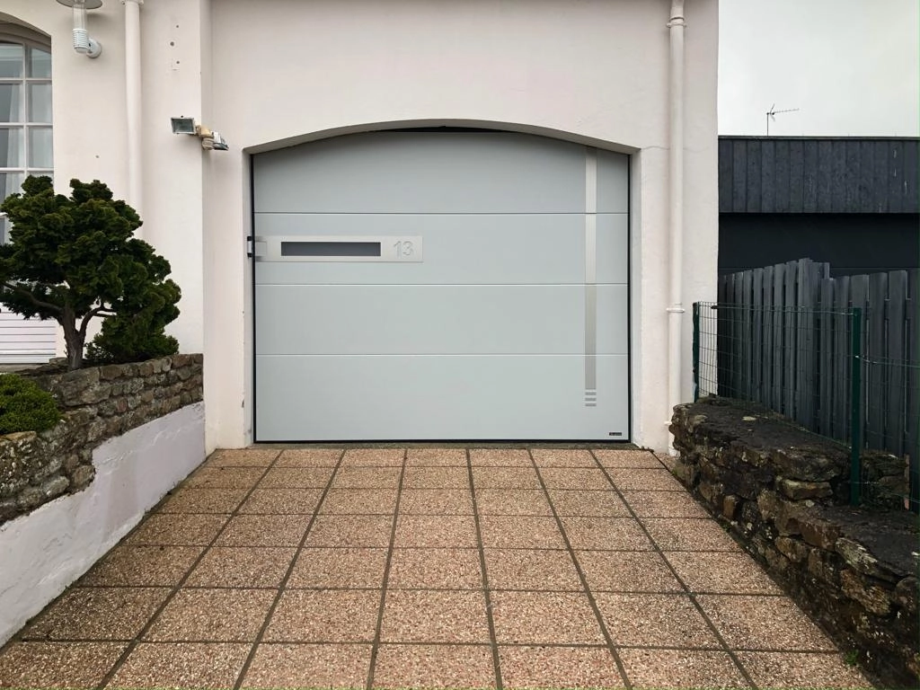 Porte garage sectionnelle grise sous arche, hublot numéro 13 et allée gravier lavé