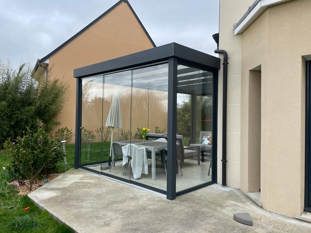 Véranda verre anthracite sur terrasse béton, table grise et parasol fermé, pelouse