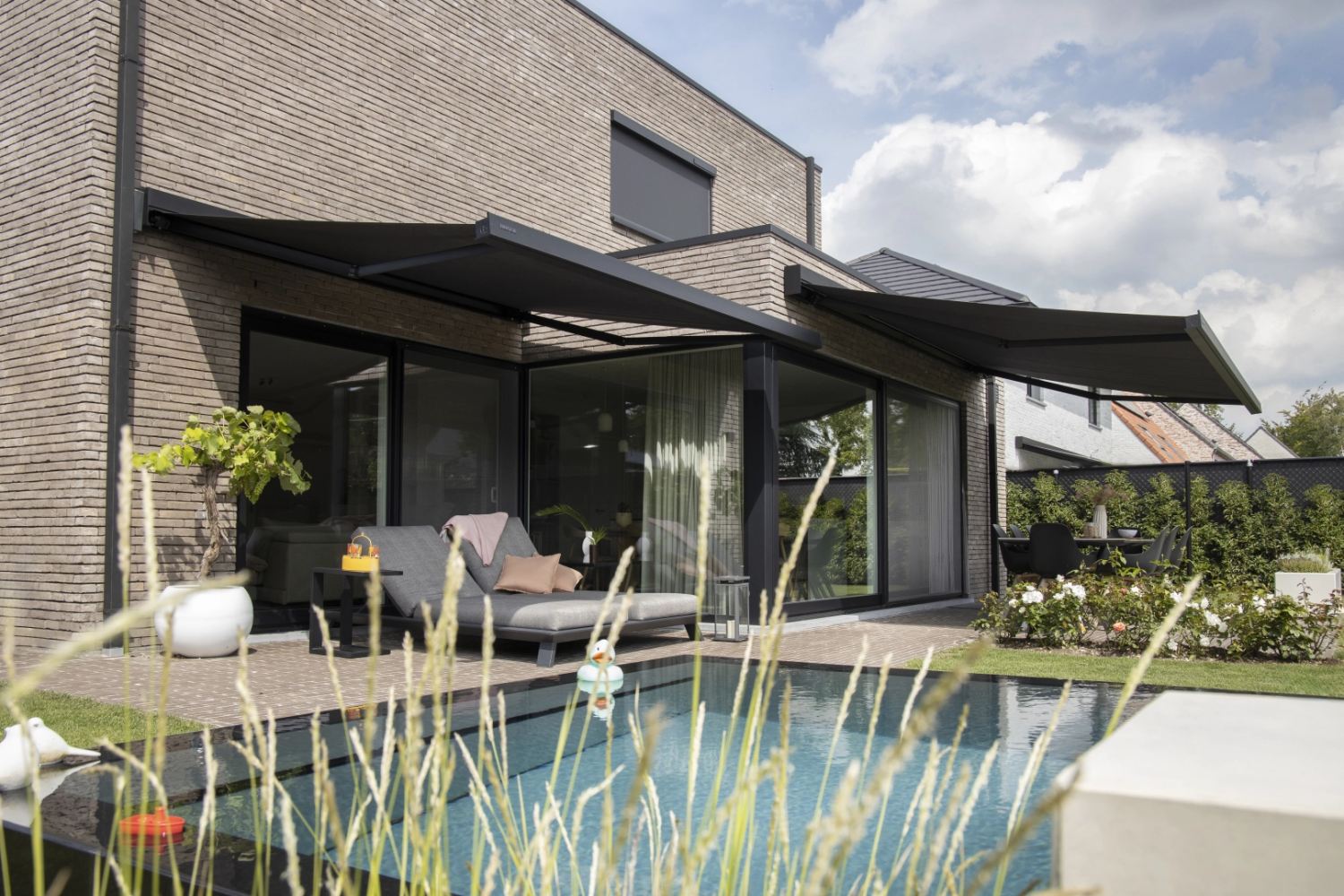 Deux stores bannes gris sur façade briques, piscine, chaises longues et table