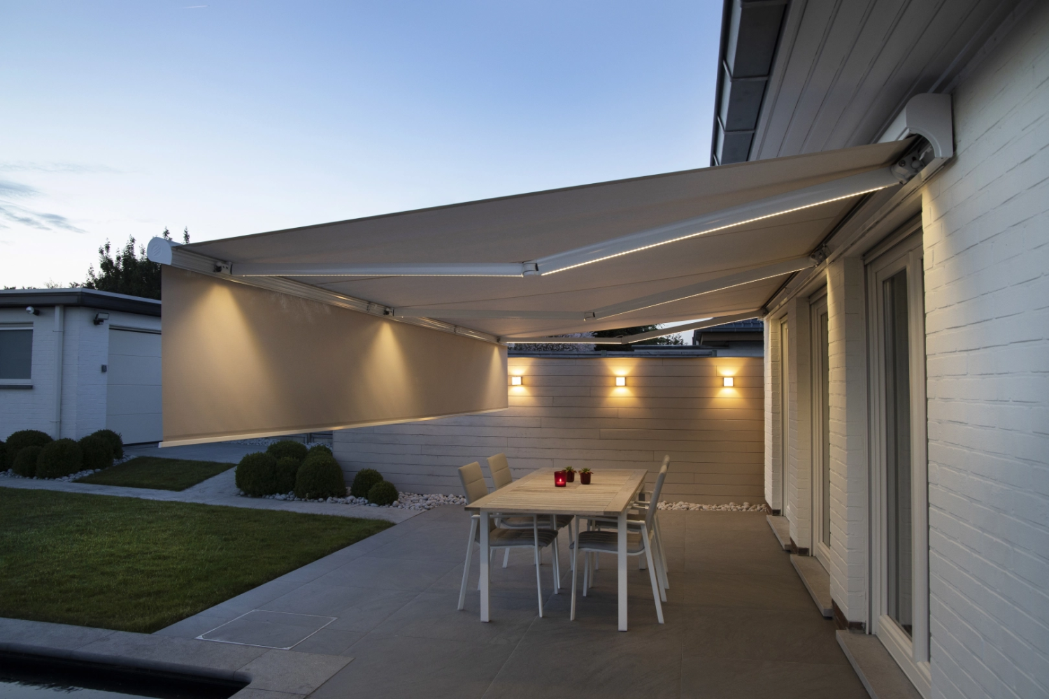 Store banne beige LED et lambrequin vertical sur terrasse crépuscule, table blanche
