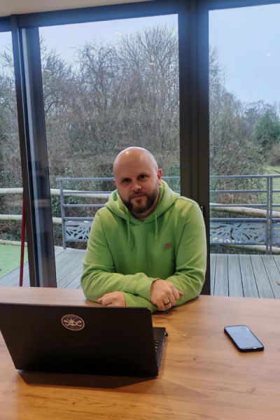 Métreur Éric en sweat vert fluo, bras croisés sur table avec ordinateur devant baie vitrée