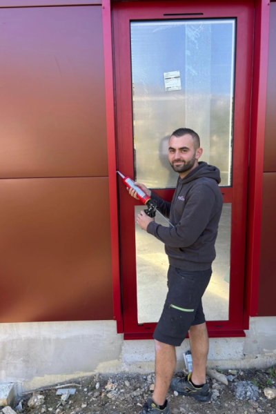 Poseur Thomas souriant appliquant mastic au pistolet sur cadre rouge d'une porte vitrée