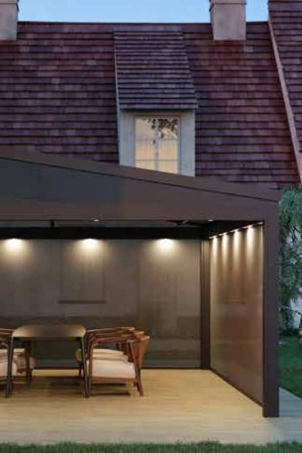Pergola Brustor anthracite éclairée le soir, stores latéraux gris et table en bois