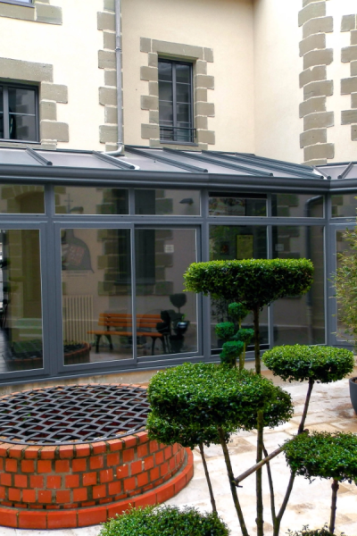 Véranda aluminium gris avec toit verre, maison pierre, puits brique et topiaires ronds